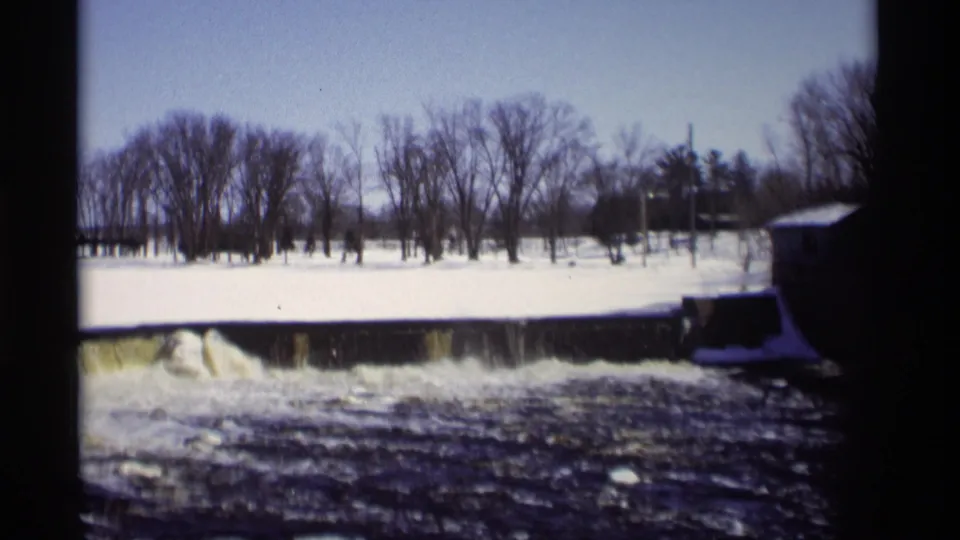 Vintage 8mm view of a winter dam in Madison, Wisconsin, with water dropping into rapids beneath a snow-covered bank.