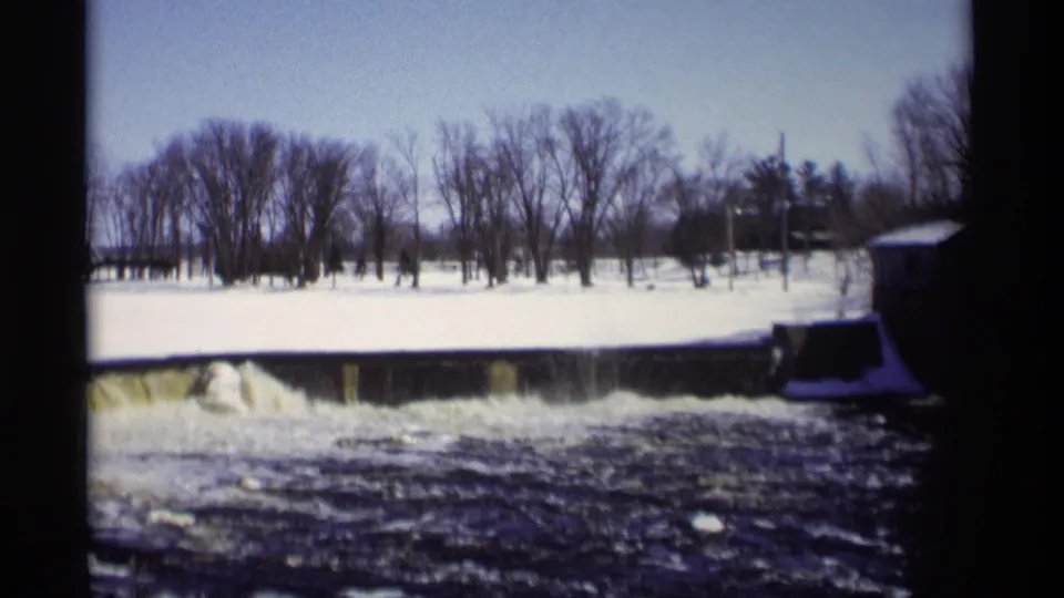 End frame of the 1966 Madison, Wisconsin winter dam scene, with a snowy bank, bare trees, and fast dark water below the spillway.