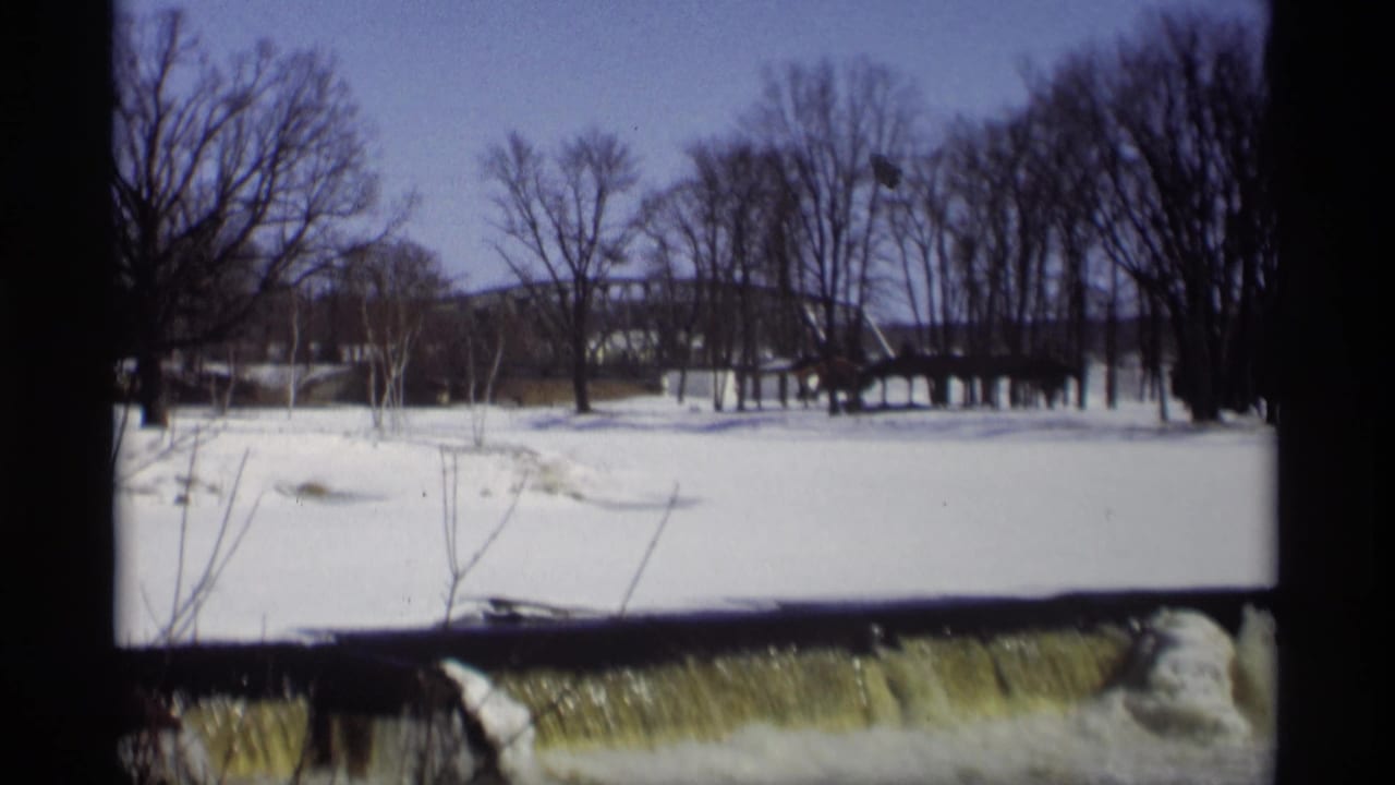 Snowy field with leafless trees, a distant bridge, and water flowing over a low dam.