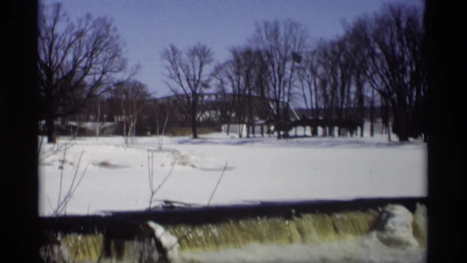 1966 Madison, Wisconsin winter view with snow, bare trees, a distant bridge, and a stream spilling over a low dam.