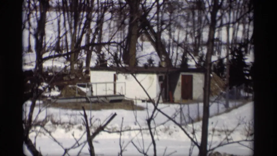 1966 Madison, Wisconsin home-movie frame showing a white shed behind leafless trees and patchy melting snow.