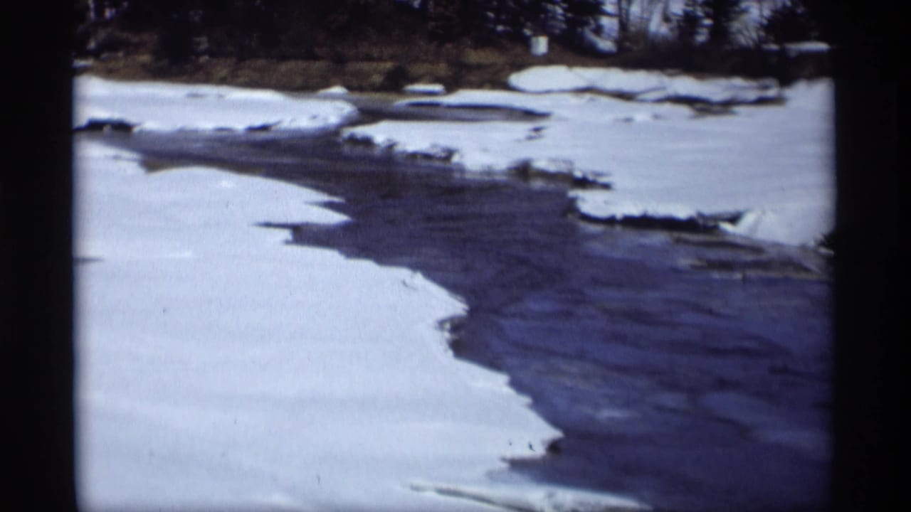 1966 Madison, Wisconsin home-movie frame showing a dark river channel cutting through snow and ice along a bare winter shoreline.