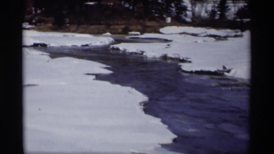 1960s 8mm frame of a partially frozen Madison river scene, with snowbanks framing dark water and leafless trees in the background.