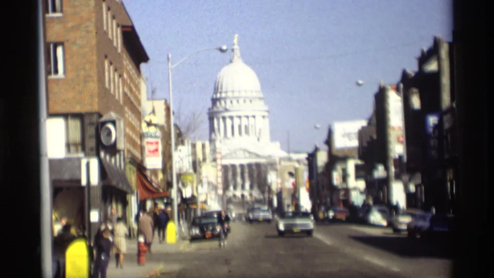 1966 Madison, Wisconsin 8mm street view looking toward the Wisconsin State Capitol, with period cars, pedestrians, storefronts, and a bicycle rider.