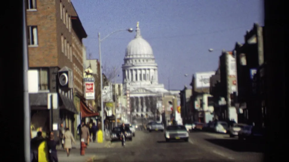 1966 Madison, Wisconsin frame showing a downtown street approach to the Wisconsin State Capitol, with moving cars, sidewalk pedestrians, and shopfront signs.