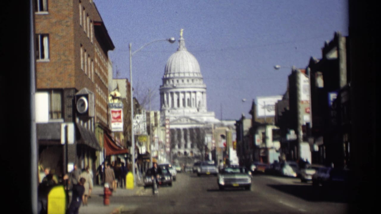 Street view toward the Wisconsin State Capitol with cars, storefronts, signs, and pedestrians.