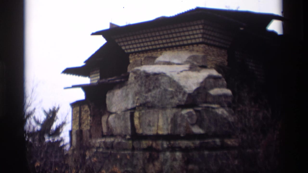 Stone building detail with a dark tiered roof behind shrubs and trees.