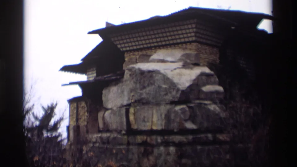 1966 Madison 8mm frame showing rough stonework, a dark tiered roof, bare shrubs, and trees in an outdoor setting.