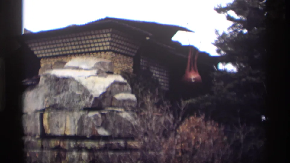 1966 Madison home-movie frame showing stonework, roof eaves, shrubs, trees, and a red hanging object near the structure.