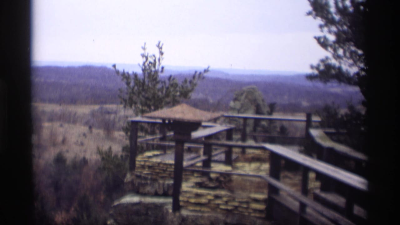 Overlook deck with wooden railing, stone wall, trees, and hazy hills beyond.
