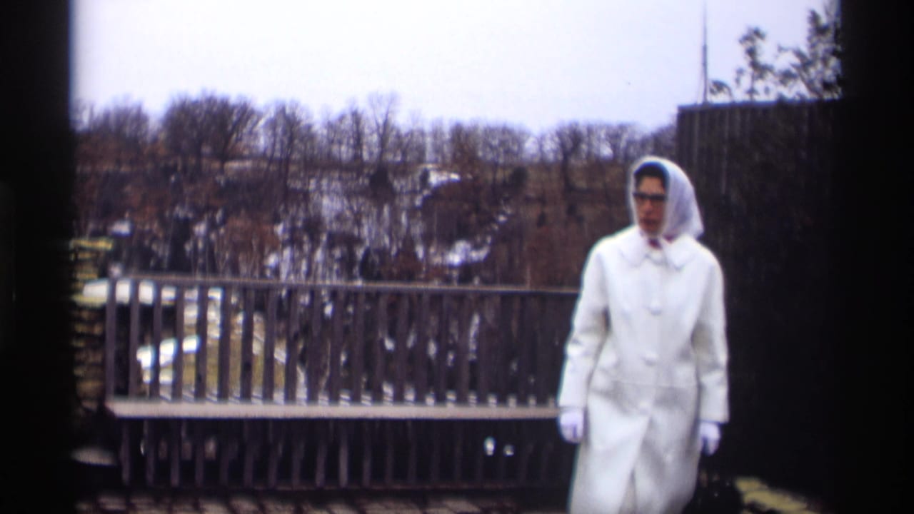 Woman in a white coat and hood walks on a stone patio near a wooden railing.