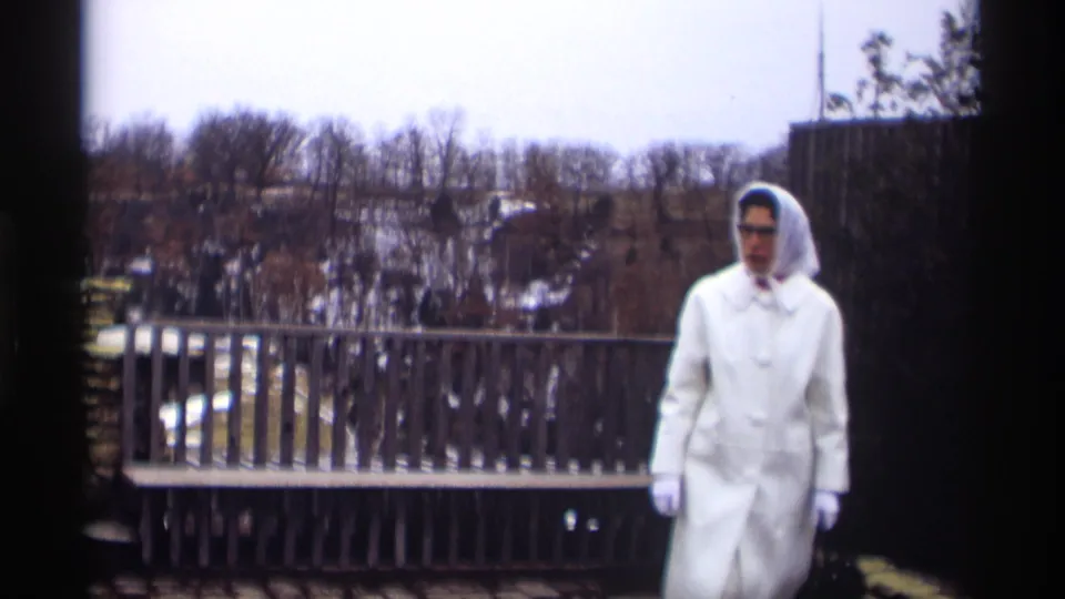 In 1966 Madison, Wisconsin footage, a woman in white winter clothing walks near a railing with bare trees and snow beyond.