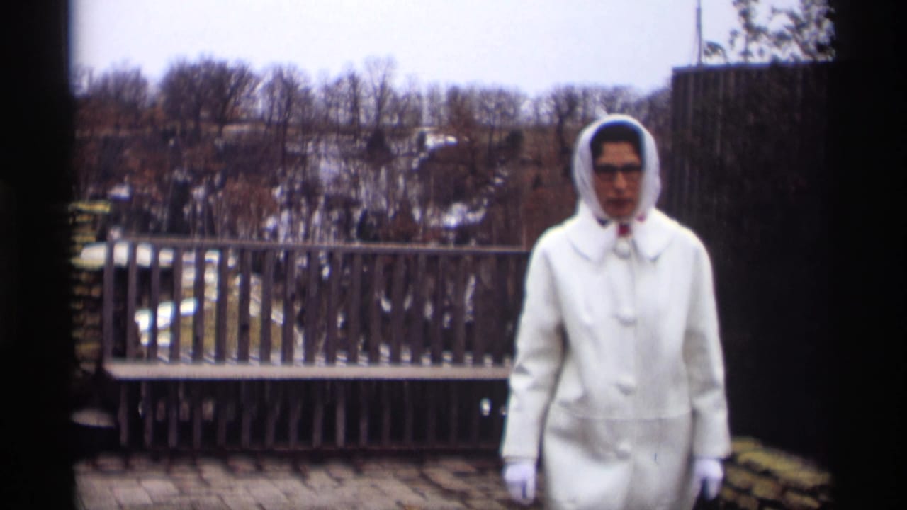 Woman in white winter clothing stands closer to the camera near a wooden railing.