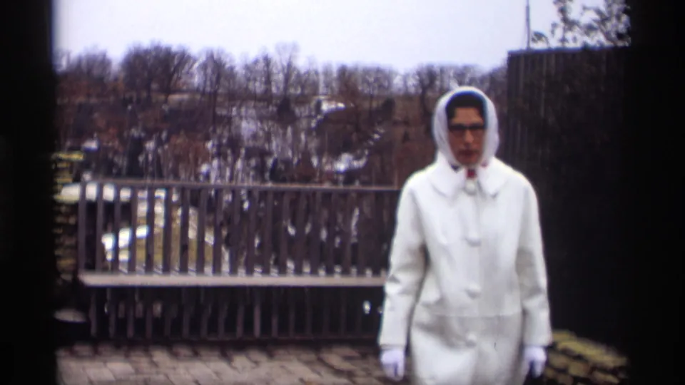 The woman in a white coat, hood, gloves, and glasses appears closer to camera on the Madison patio in 1966 home-movie footage.