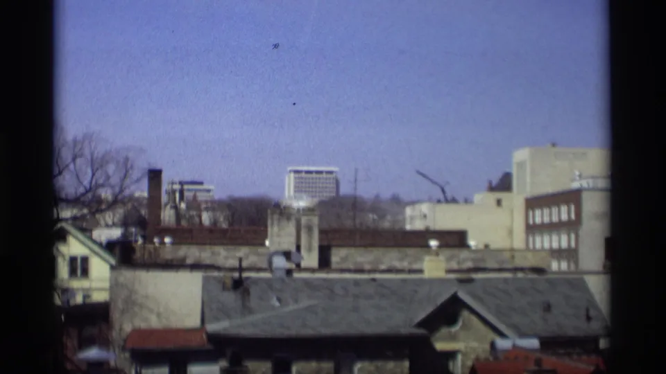 Madison, Wisconsin skyline frame showing rooftops, chimneys, pale buildings, a distant high-rise, and clear sky in 1966 footage.