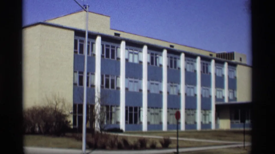 Madison 1966 home-movie frame of the columned blue-and-white building, with a stop sign, shrubs, and clear sky visible.