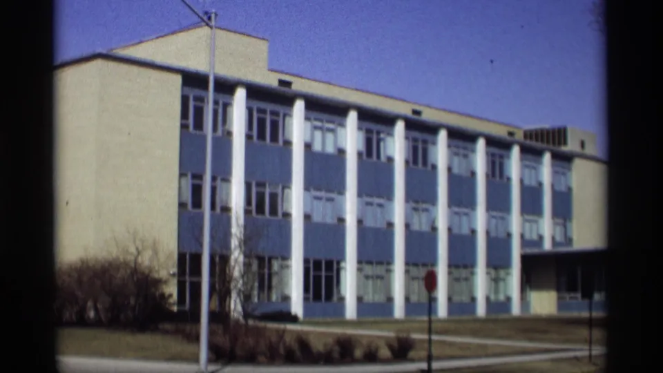 1966 Madison home-movie frame showing a blue-and-white modern building, white columns, lawn, sidewalk, and stop sign.
