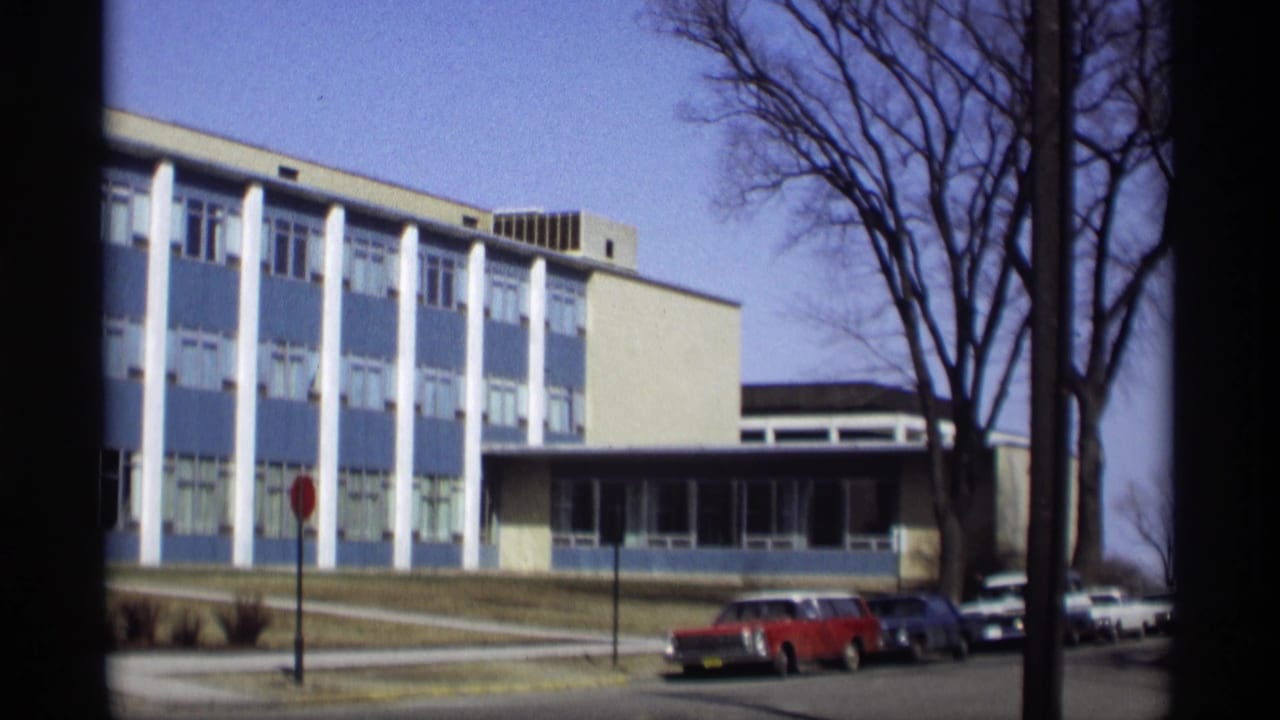 1966 Madison street view with the blue-and-white building, bare trees, sidewalk, stop sign, and parked 1960s cars.