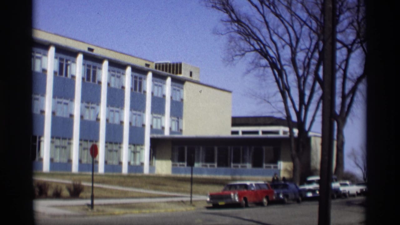 School building with blue panels, white columns, bare trees, and parked cars.