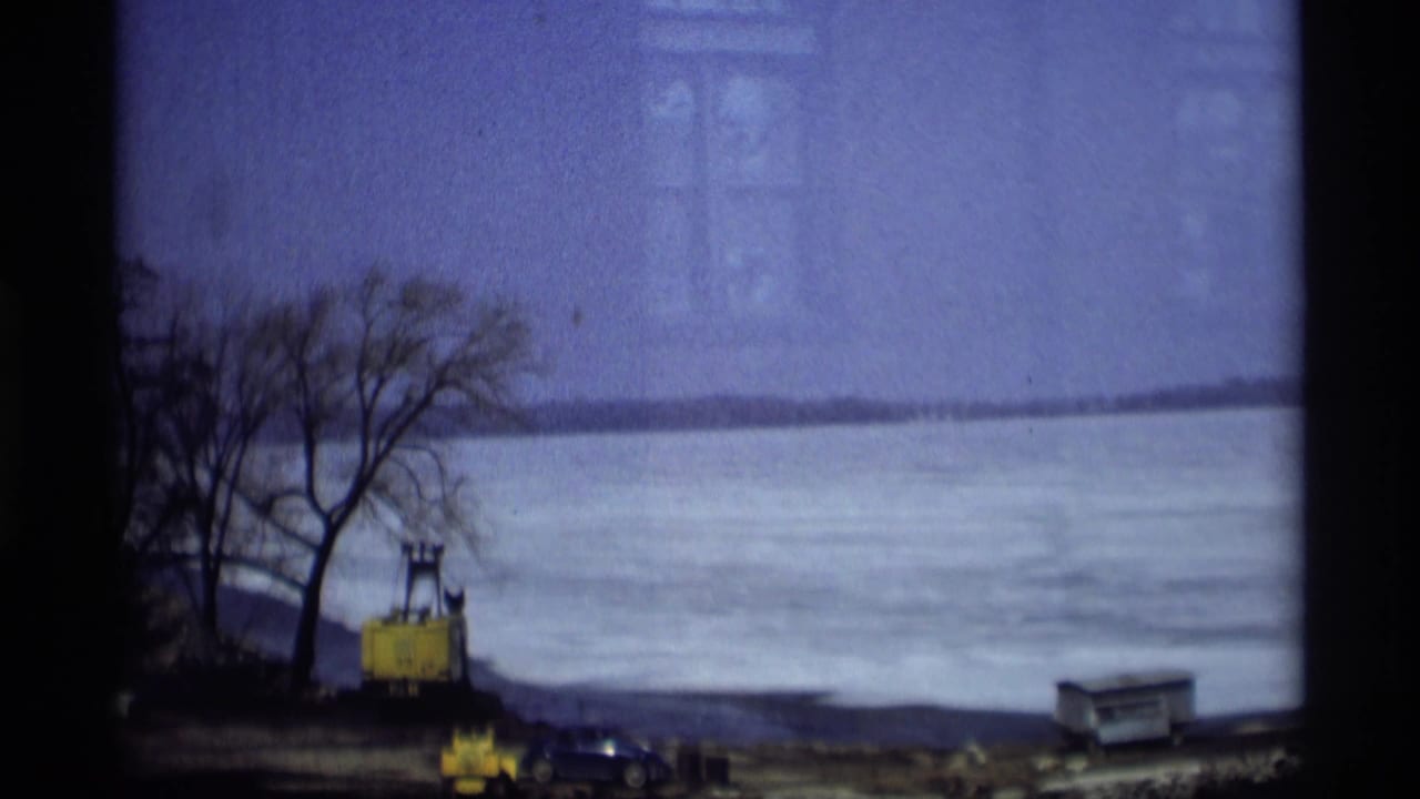 Frozen waterfront with bare trees, yellow equipment, parked cars, and distant shoreline.