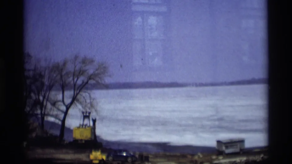 Home-movie frame of a Madison lakeshore construction area, with yellow equipment beside bare trees and cars facing an ice-covered lake.