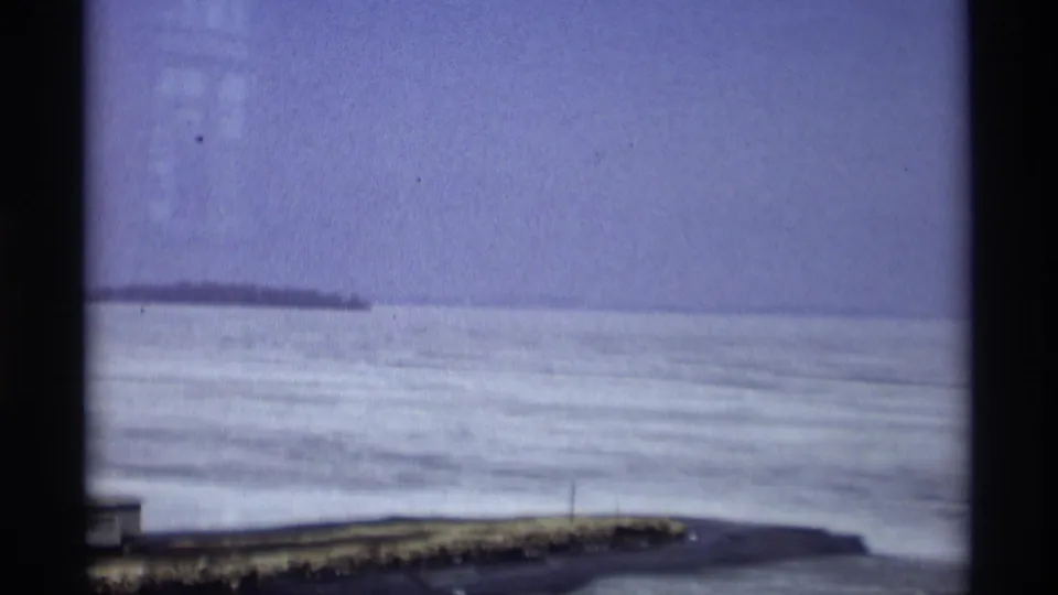 Wide Madison waterfront frame showing a frozen lake surface, low shoreline, distant land or pier, and pale purple-blue sky from the 8mm scan.