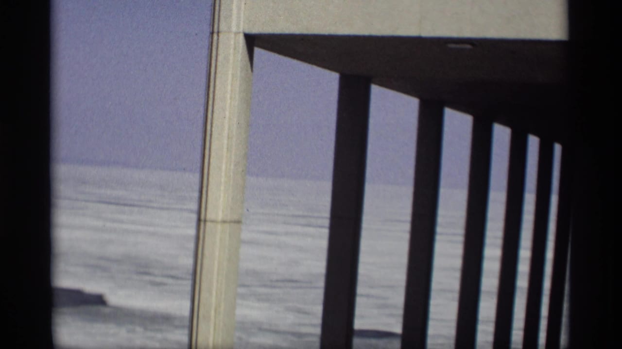 Modernist columns stand in front of calm water and pale sky.