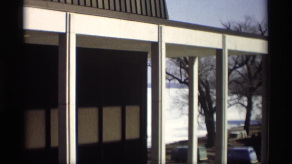 1966 8mm view of a modernist facade in Madison with glass bays, white columns, bare trees, and cars near the waterfront.