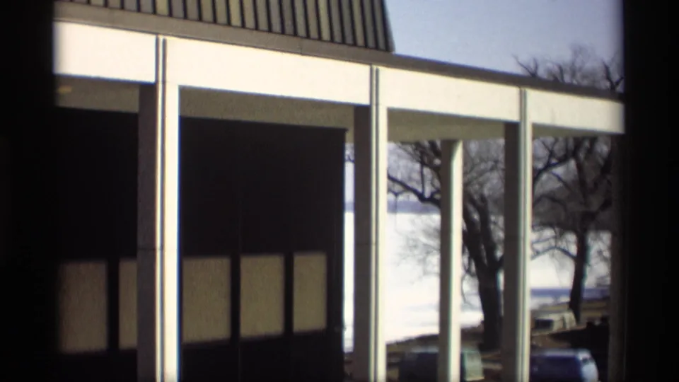 Madison 1966 home-movie frame showing the lakeside building exterior, dark window panels, white supports, bare trees, and cars.