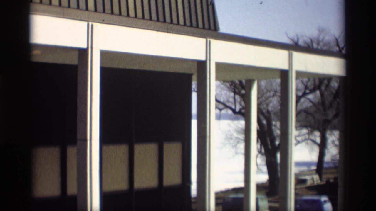 1966 Madison, Wisconsin home-movie frame showing a modern lakeside house exterior with tall windows, white supports, and bare trees near the water.