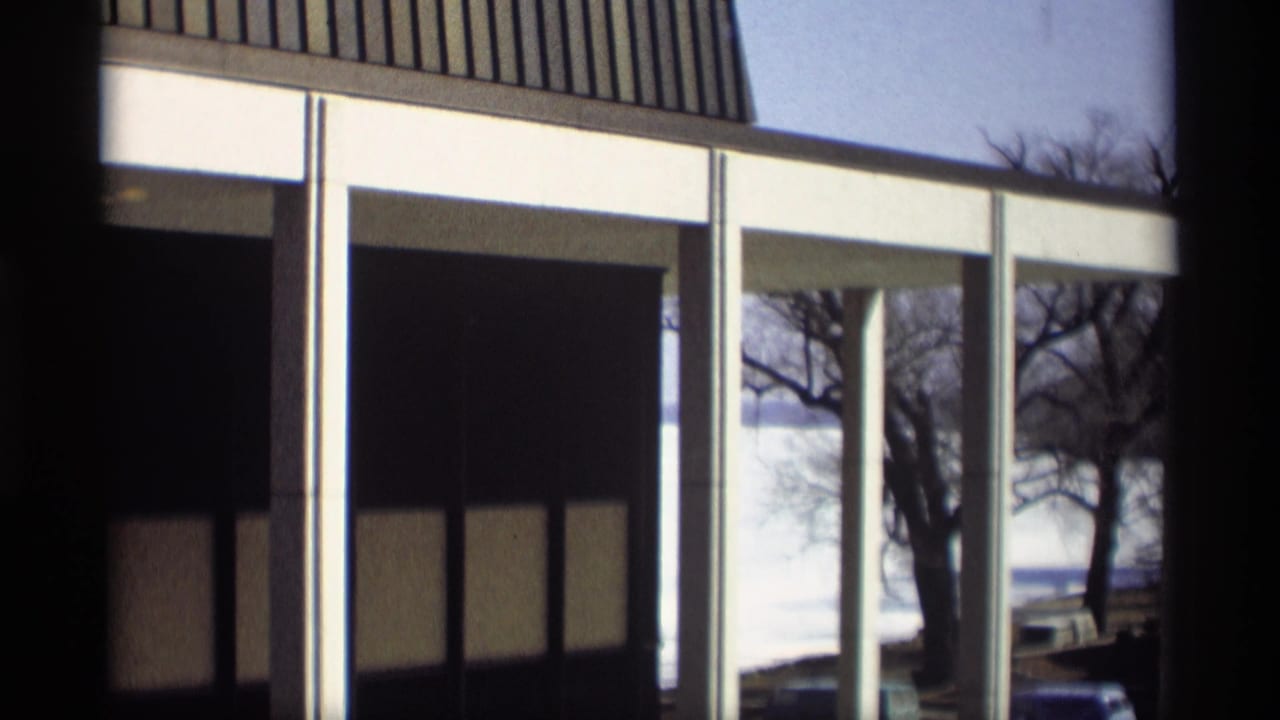 1966 Madison, Wisconsin frame holding on the lakeside house, with white columns, dark window panels, bare trees, and pale water visible beyond.