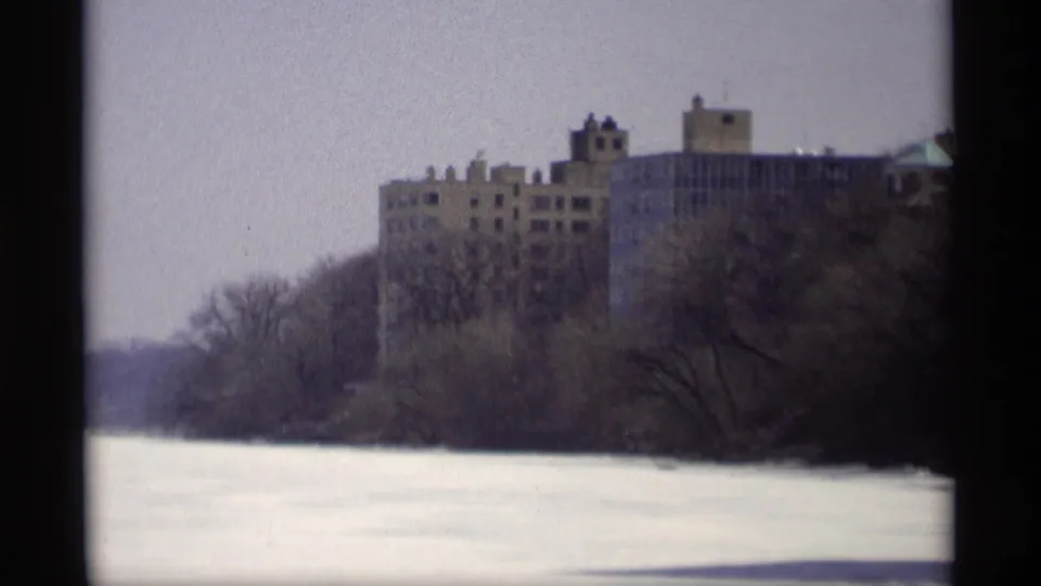 Madison, Wisconsin 1966 frame with a large brown building partly screened by winter trees beyond a frozen foreground.