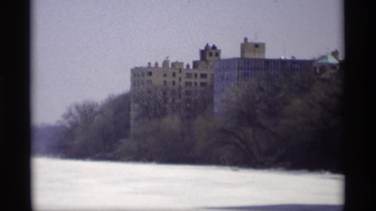 1966 Madison home-movie frame showing a distant large building across a frozen waterway with leafless trees in the foreground.