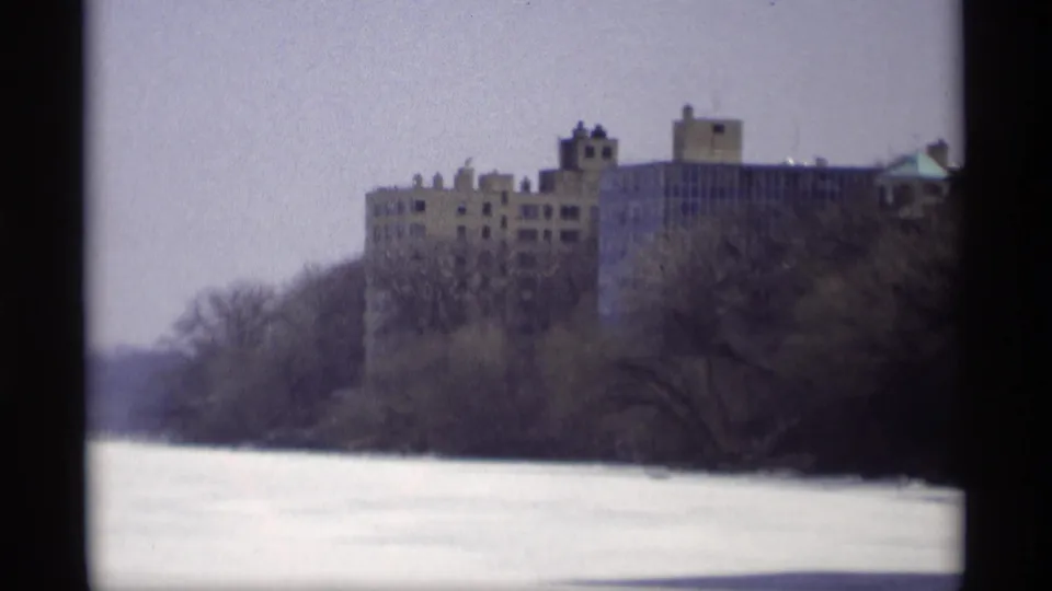 1966 Madison home-movie frame showing a distant large building across a frozen waterway with leafless trees in the foreground.