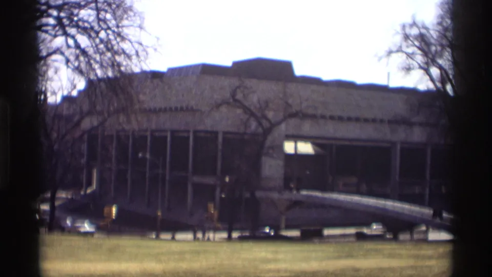 1966 Madison 8mm frame showing a broad modernist building facade, winter trees, a lawn, and parked cars near the street.