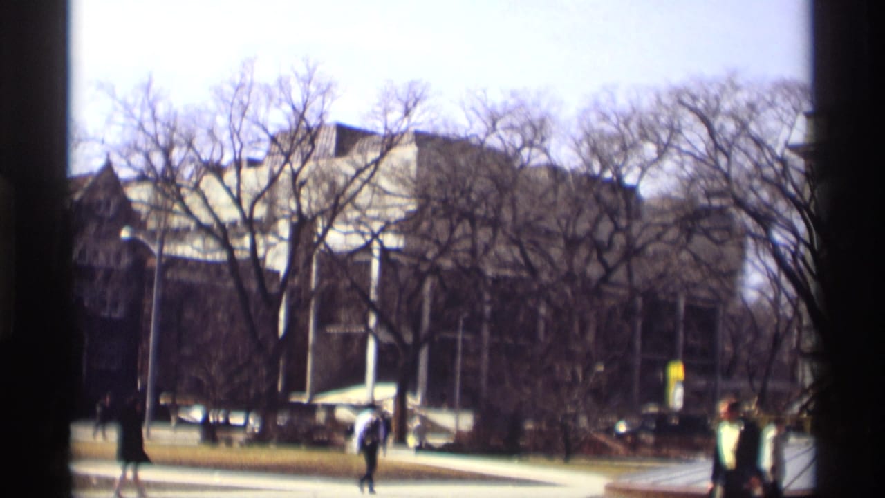 Pedestrians cross near leafless trees and a large masonry building.