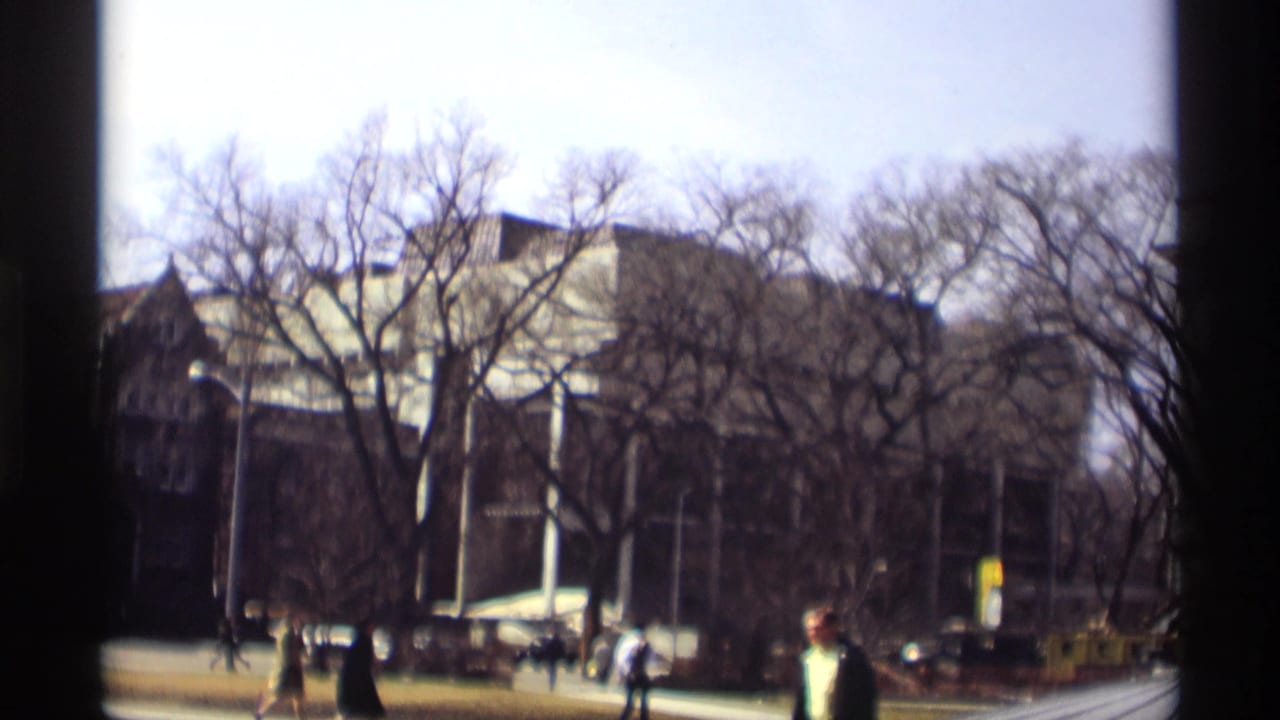 People walk along a city street with trees and a large building behind them.