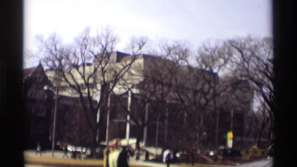 Soft 8mm frame of a Madison street with pedestrians, bare trees, cars, and a large institutional building in 1966.