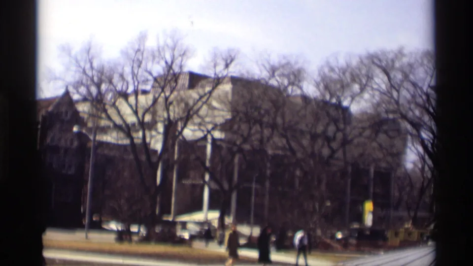 End frame of the Madison street scene with distant pedestrians, parked cars, bare trees, and a large masonry building.