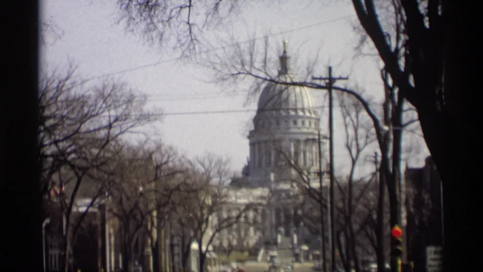 1966 home-movie frame of the Wisconsin State Capitol in Madison, viewed from a moving vehicle through winter trees.
