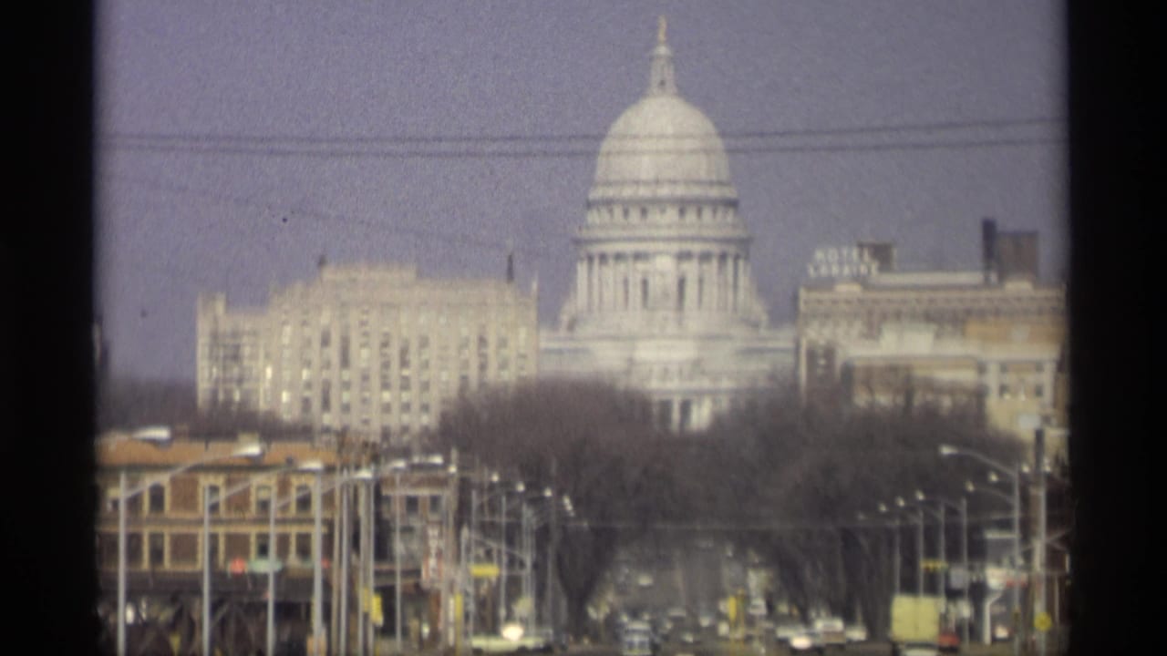 1966 Madison view of the Wisconsin State Capitol dome rising behind downtown buildings, bare trees, streetlights, and overhead wires.