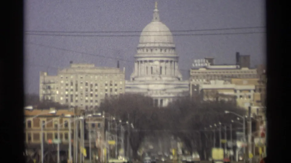 Madison, Wisconsin capitol dome seen from a distant street view with older buildings, bare trees, utility lines, and soft 8mm color.