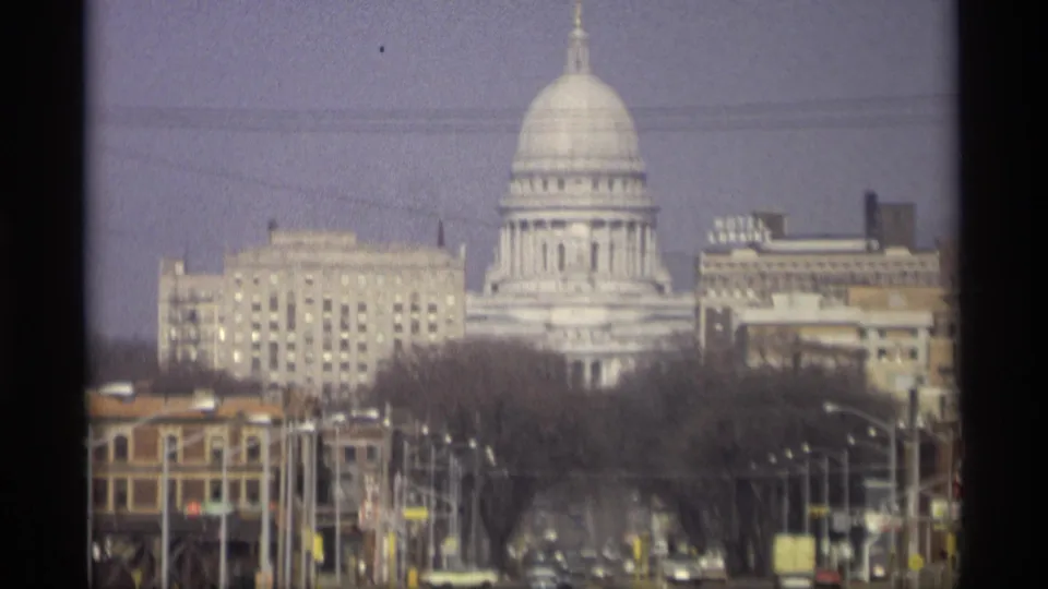 1966 home-movie frame of the Wisconsin State Capitol above Madison streets, with leafless trees, downtown facades, streetlights, and visible film softness.