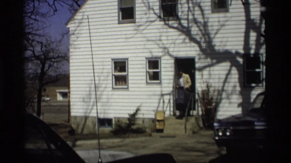 Residential exterior in Madison, Wisconsin, with a man at the doorway of a white-sided house and a suitcase near the steps.