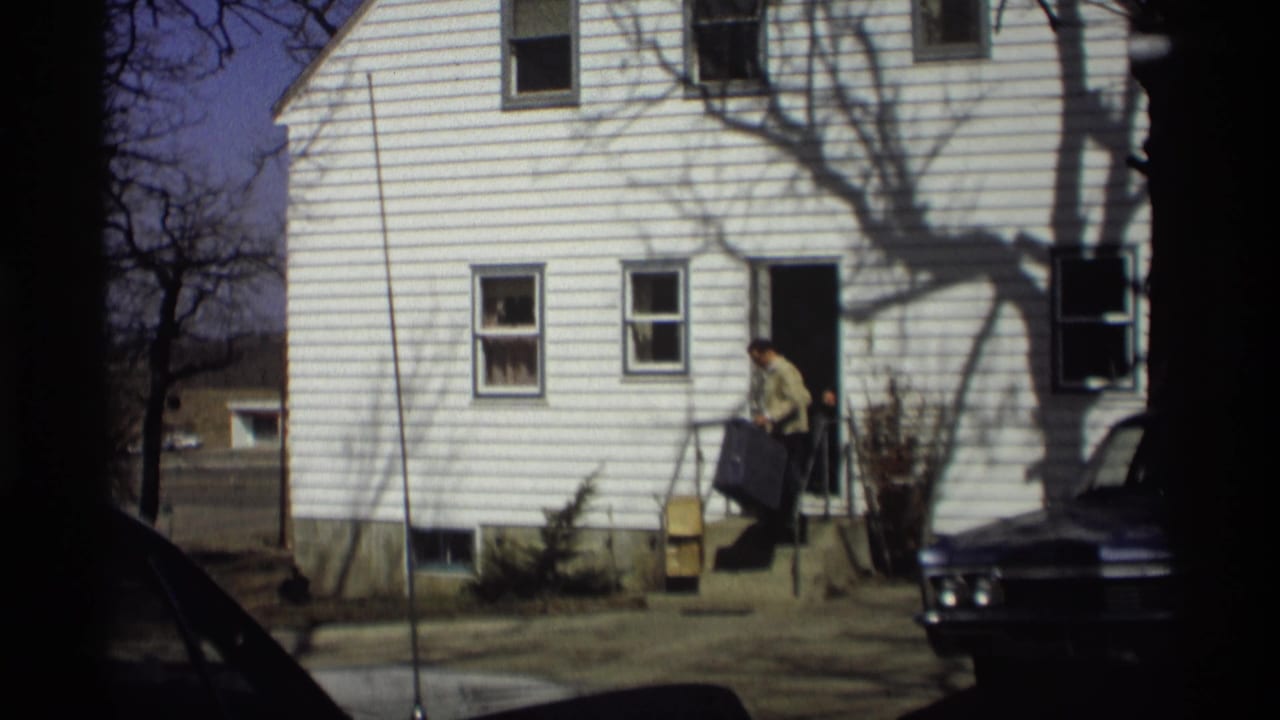 In 1966 Madison home-movie footage, a man exits a white clapboard house with a dark suitcase near a parked car.