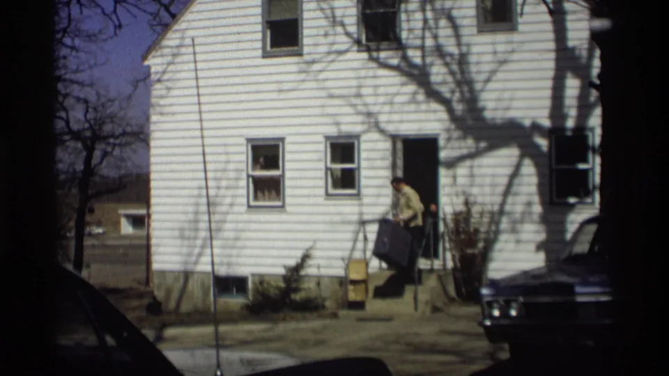 In 1966 Madison home-movie footage, a man exits a white clapboard house with a dark suitcase near a parked car.