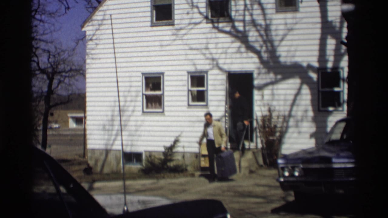 Man with suitcase stands outside a white house while another person appears in the doorway.