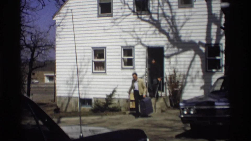 A man carrying luggage pauses outside a white Madison house as another adult stands in the doorway in 1966 footage.