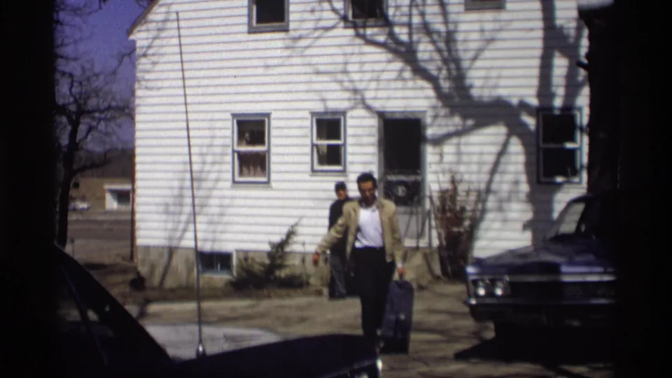 The 1966 home movie shows the man closer to camera with his suitcase, framed by the white house and a parked car.