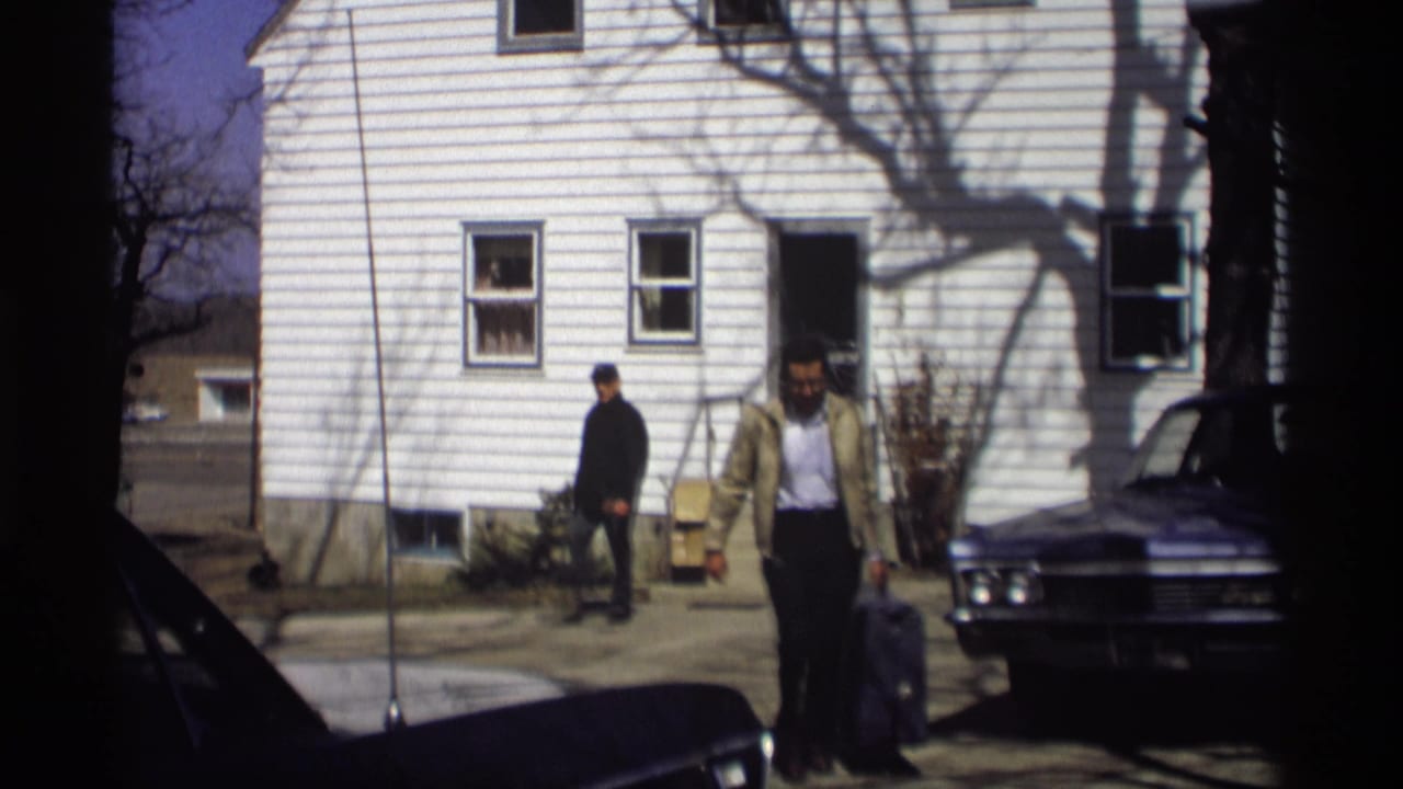 Man with a suitcase walks from a white-sided house toward a parked dark sedan.